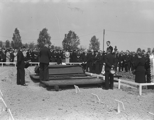 Eine Gruppe von Menschen steht um einen schwarz-weißen Sarg auf einem Friedhof, umgeben von einem Zaun, Bäumen, Pfählen und einem klaren Himmel.