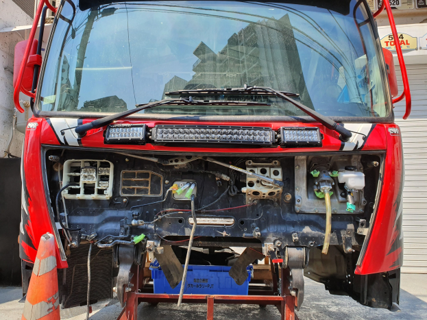 Ein roter Lastwagen, der vor einem Geb\303\244ude mit einem Verkehrskegel links und einem Rollladen rechts geparkt ist, mit Schildern mit Text und Zahlen im Hintergrund.