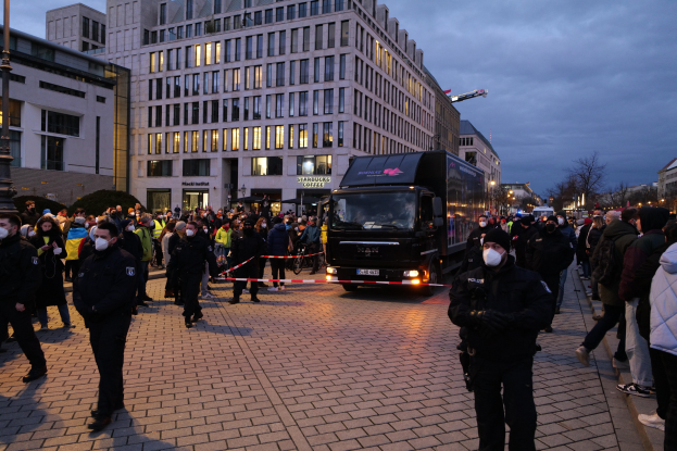 Eine Gruppe von Menschen steht in der Nähe eines Lastwagens auf einer Straße mit Gebäuden, Bäumen und Laternen im Hintergrund, einige tragen Mützen und Masken, mit einem Band und einer Stange im Vordergrund.