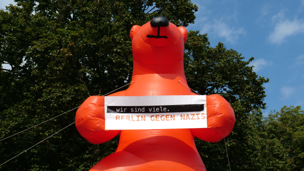 Ein aufblasbarer Bär hält ein Schild mit der Aufschrift "Berlin gegen Nazis" vor einem Hintergrund aus Bäumen und Wolken.