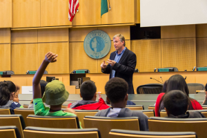 Ein Mann in einem schwarzen Blazer, blauer Hemd und schwarzen Hosen steht vor einer Klasse und h├Ąlt ein Mikrofon in der Hand, mit Kindern, die aufmerksam zuh├Âren, eine h├Âlzerne Wand mit einem Logo und zwei Flaggen dahinter und ein weißer Bildschirm rechts.