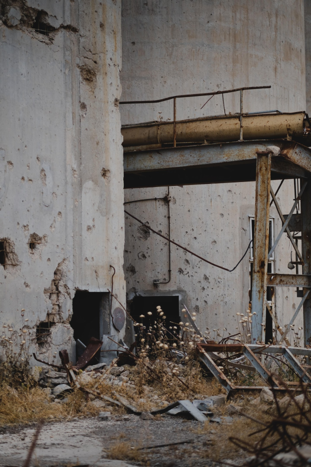 Altes Gebäude mit bröckelnden Wänden, kaputten Fenstern und einem zentralen Metallgerüst, umgeben von Pflanzen, Eisenstangen und verstreutem Schutt.
