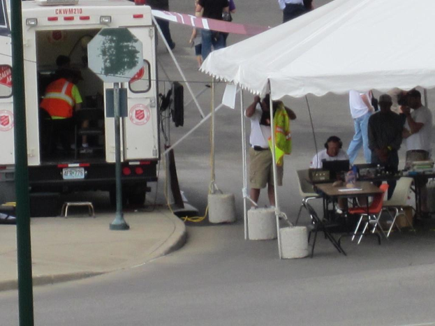 Ein Krankenwagen steht links neben einer Straße, während rechts ein Zelt eine Gruppe von Menschen schützt, bei der ein Tisch und ein Stuhl in der Nähe stehen.
