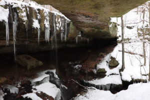 Ein kleiner Wasserfall ergießt sich über eine steinige Klippe in einem schneebedeckten Wald, mit Eiszapfen an den Felsen und schneebedeckten Bäumen.