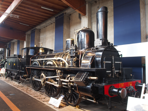 Alte Dampflokomotive im Museum ausgestellt, umgeben von Eisenbahnschienen und informativen Tafeln, mit einer Wand und beleuchteter Decke im Hintergrund.