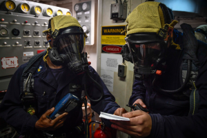 Zwei Männer in Gasmasken und Uniformen stehen nebeneinander vor einer Steuerkonsole, jeder hält ein Objekt, mit Messgeräten und einem Feuerlöscher im Hintergrund.