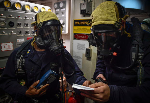 Zwei Männer in Gasmasken und Uniformen stehen nebeneinander vor einer Steuerkonsole, jeder hält ein Objekt, mit Messgeräten und einem Feuerlöscher im Hintergrund.