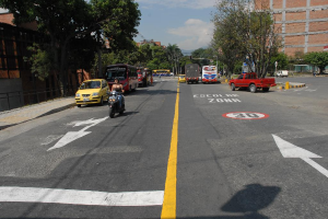 Eine Straßenszene mit Gebäuden, Bäumen, fahrenden Fahrzeugen, Verkehrszeichen, Verkehrsampeln, einem blauen bewölkten Himmel und Warnzeichen auf der Straße.