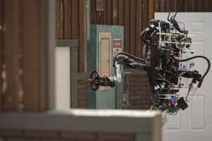 Ein humanoider Roboter mit ausgestreckten Armen und geneigtem Kopf steht vor einem Gebäude mit einer sichtbaren Tür und Wand im Hintergrund.