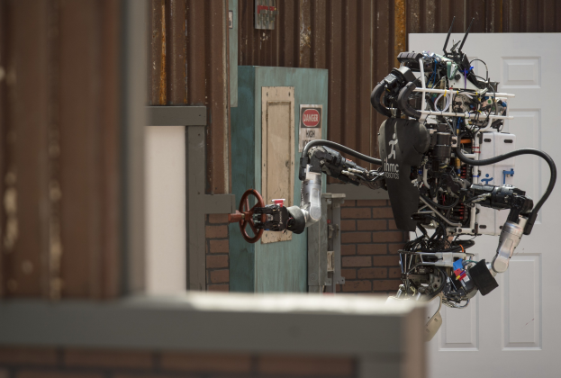 Ein humanoider Roboter mit ausgestreckten Armen und geneigtem Kopf steht vor einem Gebäude mit einer sichtbaren Tür und Wand im Hintergrund.