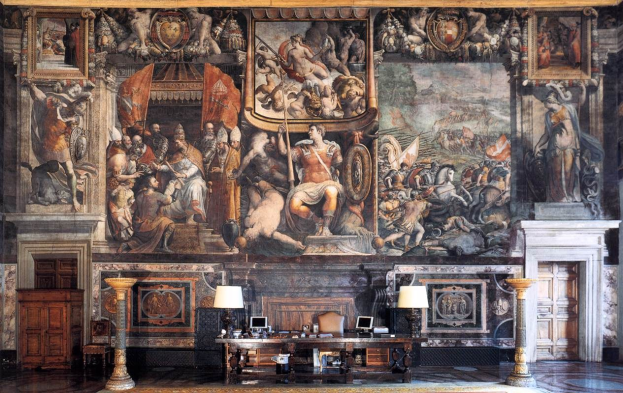 Ein großer Raum im Palazzo della Signoria in Florenz, Italien, mit einem großen Gemälde, einem Tisch mit Gegenständen, zwei Lampen, zwei Türen und einem Teppich.