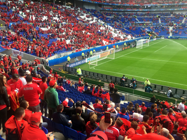 Eine große Menge an Menschen in einem Stadion schaut einem Fussballspiel zu, mit Menschen, die sitzen und stehen, Bannern, einem Torpfosten und Deckenlichtern.