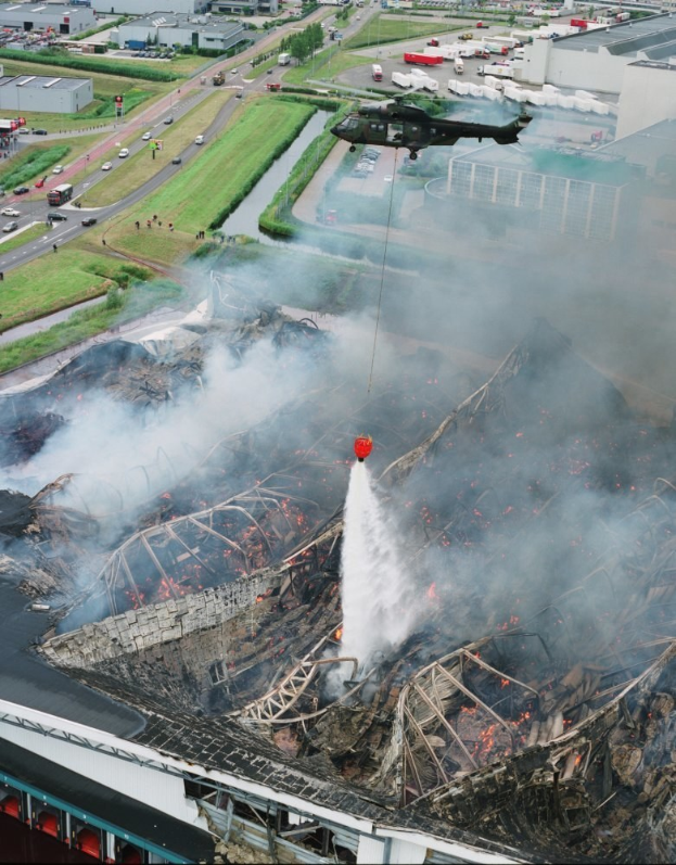Ein Hubschrauber fliegt über ein großes, durch ein Feuer zerstörtes Gebäude, aus dem Rauch aufsteigt, mit Gebäuden, Fahrzeugen und Bäumen im Hintergrund und Text am unteren Bildrand.