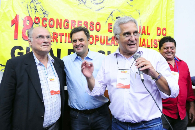 Ein Mann spricht in ein Mikrofon vor einem gelben Vorhang mit der Aufschrift "18. Kongress o", während andere Männer im Hintergrund stehen.