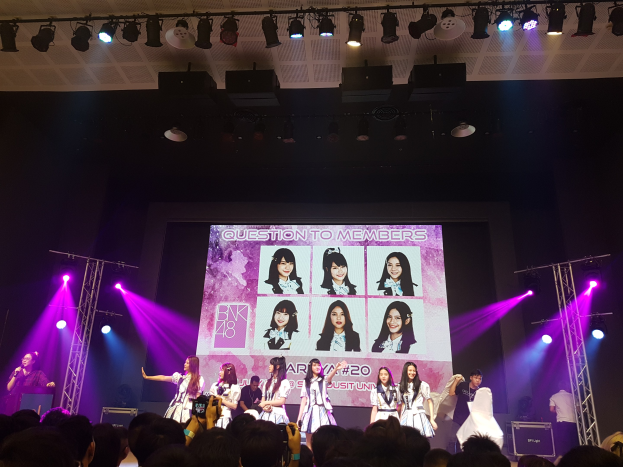 Eine Gruppe von Menschen performs auf einer hell erleuchteten Bühne mit Trägern, mit einem Sänger, der ein Mikrofon hält, und einem Musiker, der ein Instrument spielt, während eine Menge unten das Ereignis mit Kameras aufzeichnet und ein großer Bildschirm Bilder der K-Pop-Gruppe BK48 anzeigt.