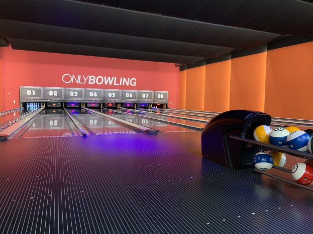 Eine Bowlingbahn mit Bowlingkugeln auf der rechten Seite, Bowlingpins in der Mitte und einer textbedeckten Wand im Hintergrund, mit der Decke darüber.
