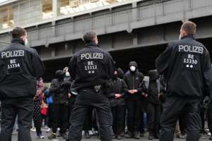 Eine Gruppe von Polizisten in Uniform steht vor einer Menge von Menschen in schwarzen Uniformen und Masken, mit einer Brücke und einem Gebäude im Hintergrund, während einer Demonstration in einer Stadt.