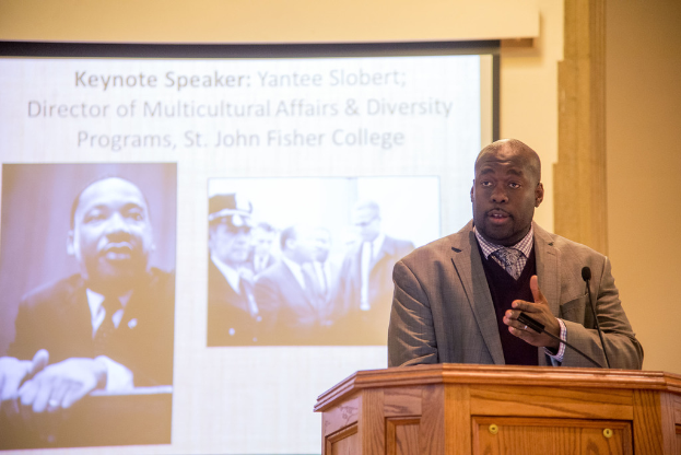 Ein Mann in Mantel, Krawatte und Hemd spricht neben einem Rednerpult auf der rechten Seite des Bildes, mit einem Projektionsschirm in der Mitte und einem Mann in Schwarz und Weiß auf der linken Seite.