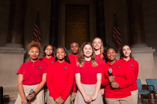 Gruppe junger Frauen in roten Shirts, die strahlend Mikrofone halten und vor Stühlen, Säulen, Fahnen und einer Wand stehen.