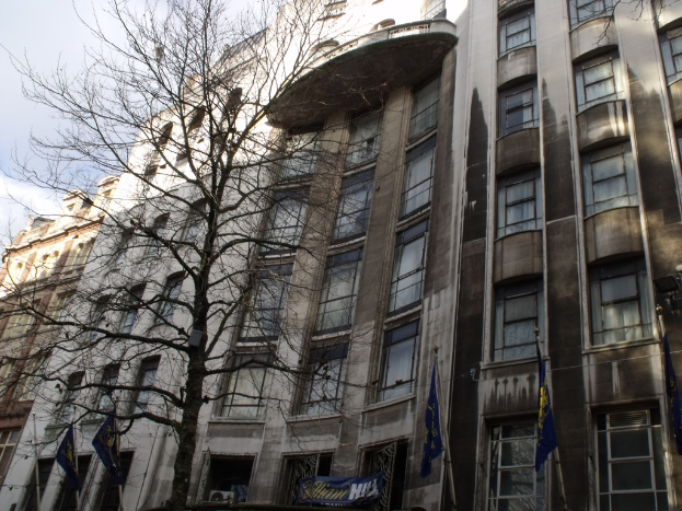 Geb├Ąude mit Fenstern, einem Banner und Fahnen an Masten unten und einem Baum mit Zweigen.