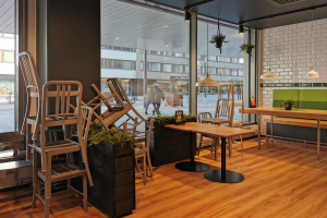 Ein Restaurant-Innenraum mit Holztischen und -stühlen, großen Fenstern mit Blick auf eine Stadtstraße mit Menschen, Gebäuden und Pfählen, umgeben von Pflanzen und Lichtern.