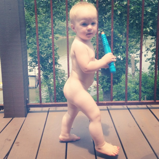 Ein Kind, das ein Objekt hält und vor einem Haus, Eisenstäben, Bäumen und Wasser steht.