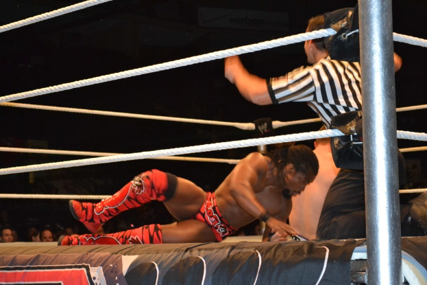 Zwei Männer kämpfen in einem Ring, einer trägt ein rotes Hemd und Schuhe, der andere Shorts, mit einem Schiedsrichter rechts daneben und einer Zuschauermenge im Hintergrund unter einem dunklen Himmel.