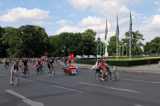 Eine Gruppe von Menschen, die auf Fahrrädern eine von Bäumen gesäumte Straße mit Laternenmasten und Fahnenmasten entlangfahren, während der Pariser Fahrradtag 2016, mit bewölktem Himmel und Fahrzeugen im Hintergrund.