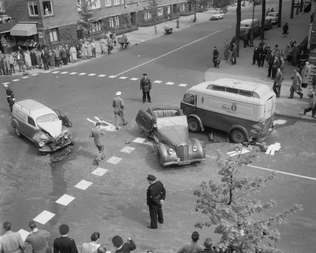 Schwarz-weißes Bild eines Autounfalls an einer Straßenkreuzung mit einem Van, anderen Fahrzeugen und Menschen in der Nähe, mit Gebäuden, Bäumen, Pfählen und Texttafeln im Hintergrund.