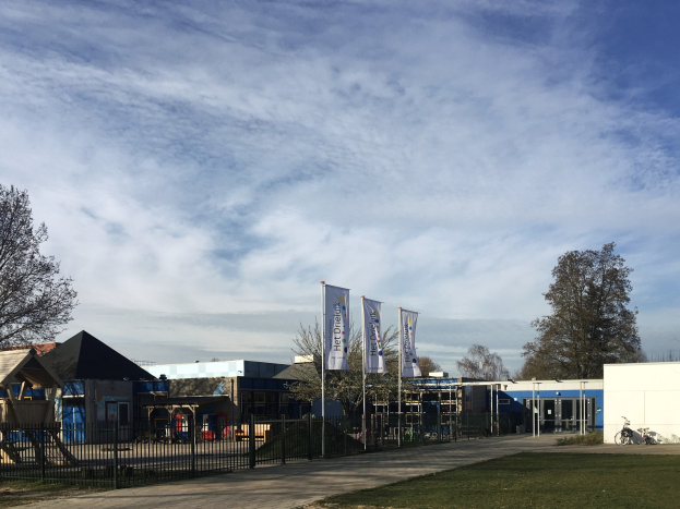 Großes Gebäude mit Metallzaun, Rasenfläche, Weg, Bäumen, Pfosten, Bannern, einem geparkten Fahrrad, nahen Häusern und einem bewölkten Himmel, identifiziert als der neue Spielplatz für Kinder.
