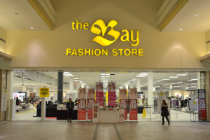Außenansicht eines Bay Fashion Stores in einem Einkaufszentrum mit Einkaufenden, Wandtafeln, Säulen, Deckenleuchten und Hintergrundauslagen.
