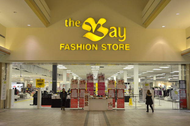 Außenansicht eines Bay Fashion Stores in einem Einkaufszentrum mit Einkaufenden, Wandtafeln, Säulen, Deckenleuchten und Hintergrundauslagen.