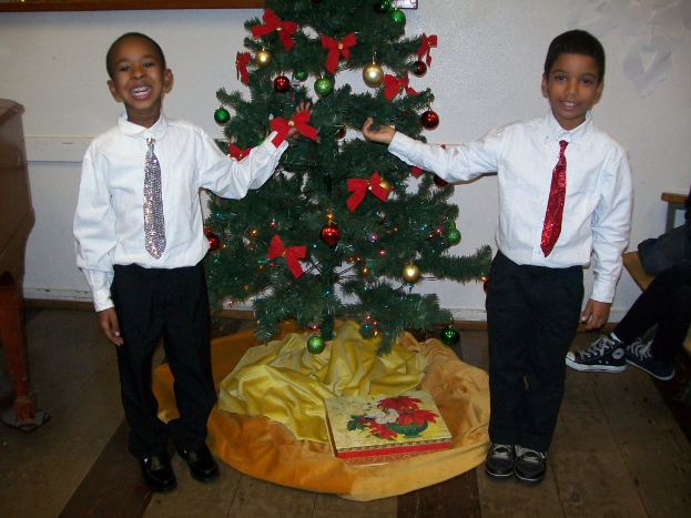 Ein geschmückter Weihnachtsbaum steht in einem Raum mit einem Holztisch links, einem Stuhl rechts und zwei Jungen in weißen Hemden, die lächeln und den Baum halten.