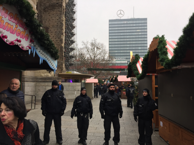 Eine Gruppe von Polizisten in schwarzen Uniformen und Mützen steht vor einem festlichen Weihnachtsmarkt in Berlin, Deutschland, mit Gebäuden, Bäumen und einem klaren blauen Himmel im Hintergrund und einer Tafel mit Schrift auf der linken Seite des Bildes.