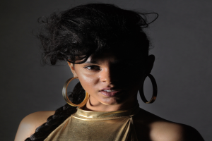 Eine Frau in einem goldenen Kleid und großen Kreolen steht selbstbewusst vor einer Wand, ihr glatter Bob-Haarschnitt und das natürliche Make-up unterstreichen ihren entschlossenen Gesichtsausdruck, während sie direkt in die Kamera blickt.