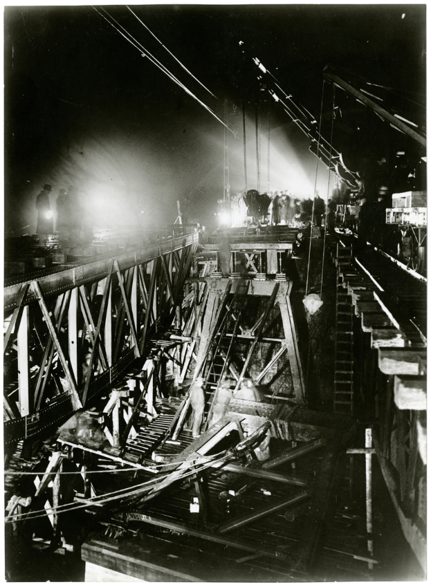 Ein Schwarz-Weiß-Foto einer Brücke im Bau, mit ein paar Menschen auf der Brücke und ein paar Lichtern, die die Gegend erhellen.