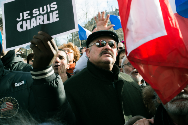Eine Gruppe von Menschen, die auf einer Straße protestieren und Fahnen und Plakate halten, mit Bäumen und dem Himmel im Hintergrund.