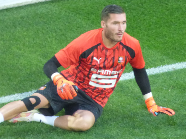 Ein Fußballer in rotem T-Shirt, schwarzen Shorts, weißen Socken und orangefarbenen Baseball-Handschuhen sitzt auf dem Boden mit Händen auf den Knien.