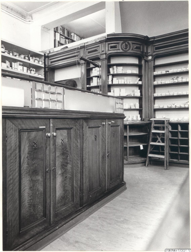 Ein Schwarz-Weiß-Foto einer Apotheken-Inneneinrichtung mit einem Tresen, Schränken, einer Leiter und Regalen, die mit verschiedenen Gegenständen gefüllt sind, mit Text oben auf dem Bild.