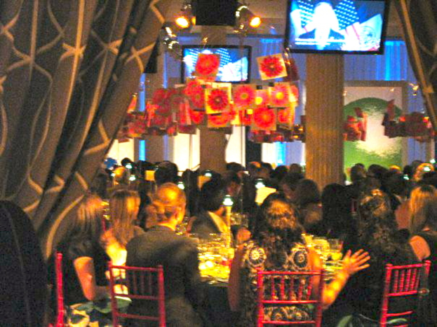 Eine Gruppe von Menschen sitzt an Tischen mit Essen, Fernsehern, Lampen, hängenden Papieren und Vorhängen in einem scheinbar festlichen Setting.