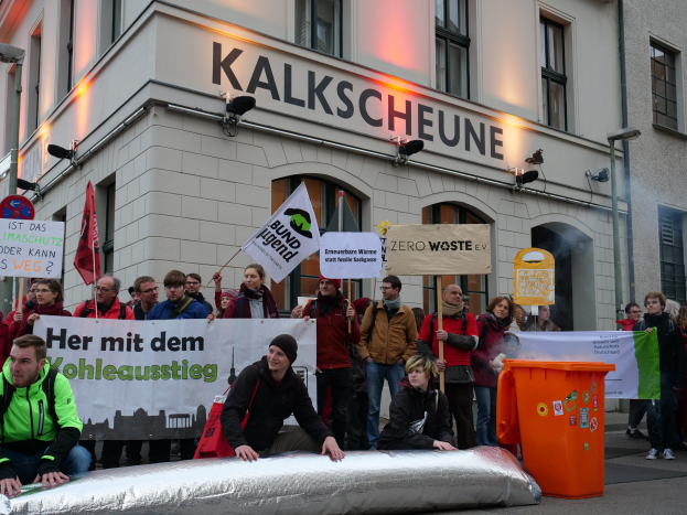 Eine Gruppe von Menschen, die vor einem Gebäude in Deutschland protestieren, mit Schildern und Plakaten, zwei Personen im Vordergrund und ein Mülleimer auf der rechten Seite.