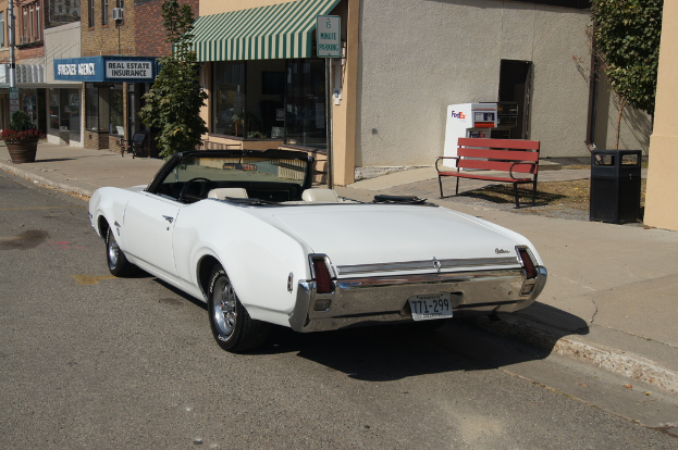 Ein weißes Auto mit dem Kennzeichen 771-299 steht auf der Straße, rechts daneben eine Bank, im Hintergrund ein Gebäude und ein Geschäft namens 'Immobilienversicherung'.