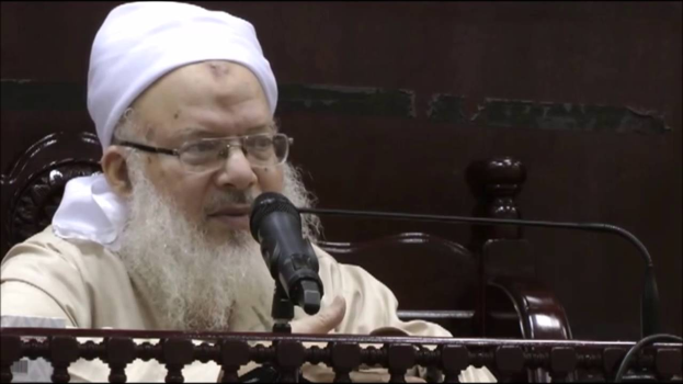 Asaduddin Owaisi, ein Mann mit weißem Bart und Turban, spricht in ein Mikrofon vor einer hölzernen Absperrung, trägt ein cremefarbenes Gewand und Brille vor dunklem Hintergrund.