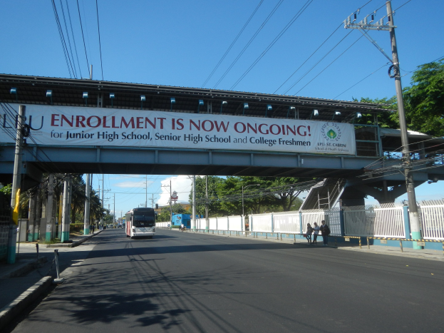 Ein Bus fährt unter einer Brücke mit einem Schild, auf dem "Jetzt Anmeldung" steht, während Fußgänger darunter gehen und Strommasten die Straße säumen, mit Bäumen und einem klaren Himmel im Hintergrund.