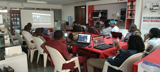 Gruppe von Menschen an einem Tisch mit Laptops und anderen Gegenständen während eines digitalen Marketing-Workshops, mit einem Banner, einem Bildschirm, einer Tür und Deckenleuchten im Hintergrund.