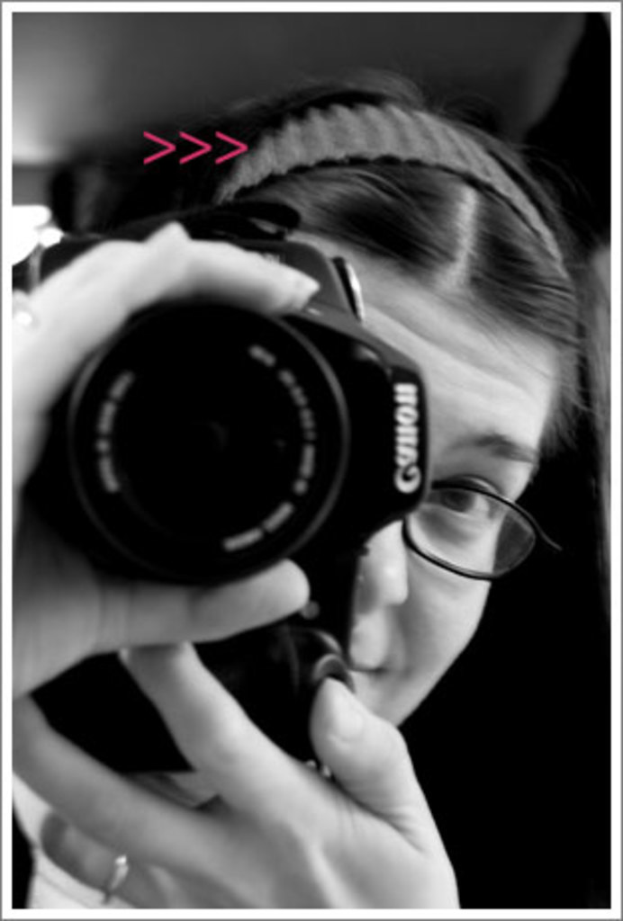 Eine Frau mit Brille, Haarband und Ring lächelt und hält eine Kamera in einem Schwarz-Weiß-Bild.