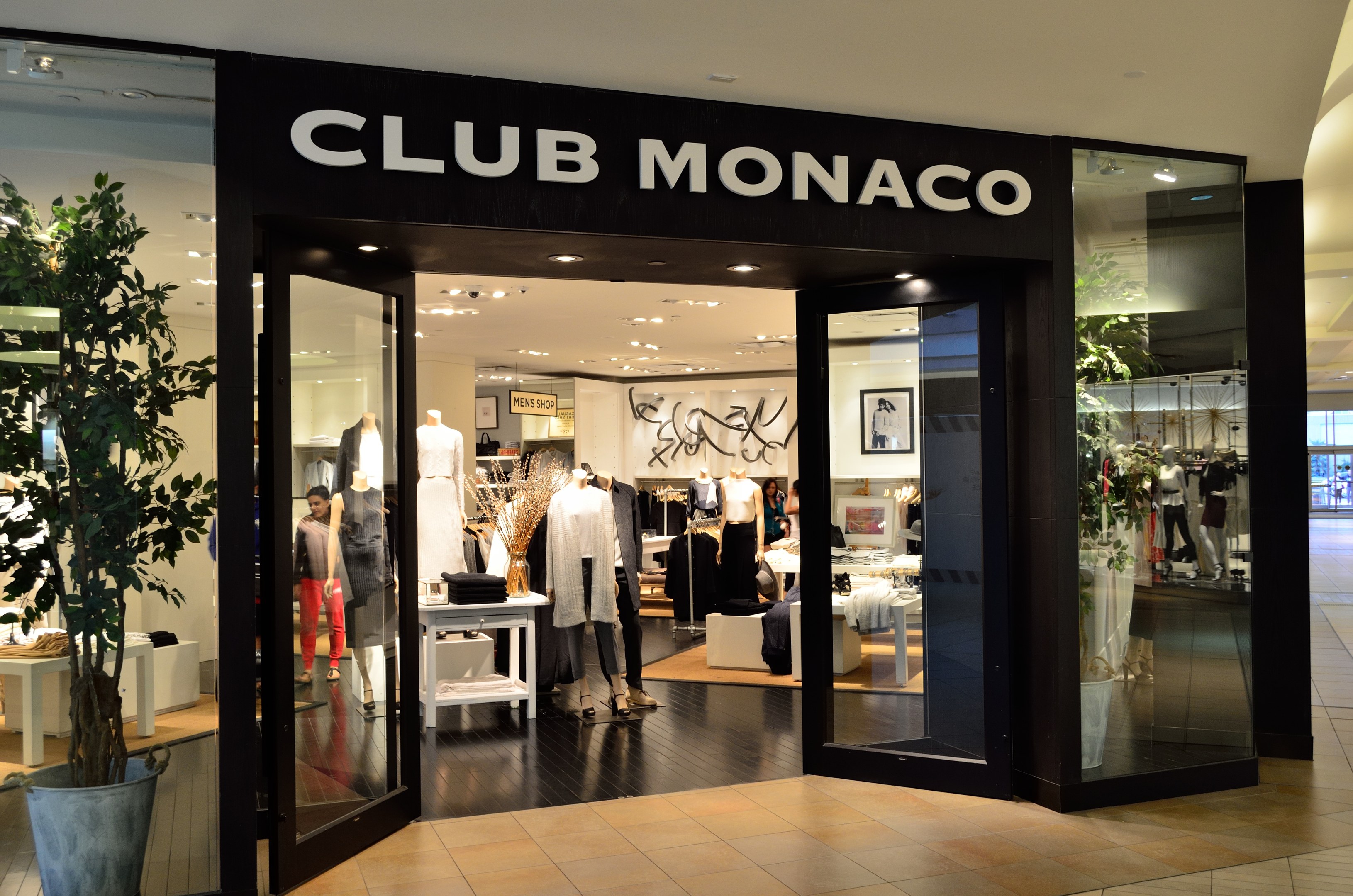 Vorderfront eines Club Monaco Einzelhandelsgeschäfts in einem Einkaufszentrum mit Glaswänden und -türen, Schaufensterpuppen in Kleidern, Auslagetischen mit Waren, einem Topf mit einer Pflanze und Deckenbeleuchtung.