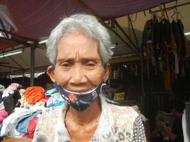 Eine ältere Frau mit Mund-Nasen-Schutz steht auf einem Markt umgeben von hängenden Klamotten, Lichtern, Ventilatoren und anderen Gegenständen, mit einer Person im Hintergrund sitzend.