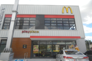 Ein McDonald's-Restaurant mit parkenden Autos davor, umgeben von Bäumen und Polen unter einem klaren blauen Himmel, mit Glaswänden, Fenstern und einem "McDonald's Playplace"-Schild, mit sichtbaren Postern und Gegenständen im Inneren.