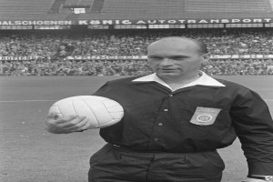 Ein Schwarz-Weiß-Foto eines Schiedsrichters auf einem Fußballfeld, der einen Ball hält, mit Zuschauern und Tafeln mit Text im Hintergrund.